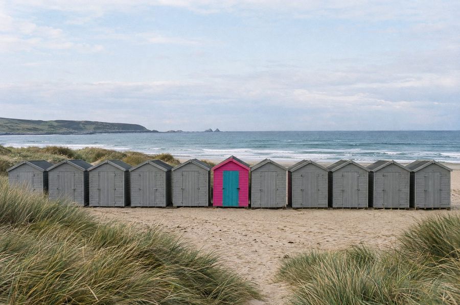 A row of identical grey beach huts with one painted pink with a teal door